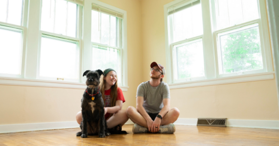 A woman and man sitting on the floor of their home with their dog
